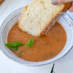 A bowl of tomato soup with a hand dipping a slice of bread in it.