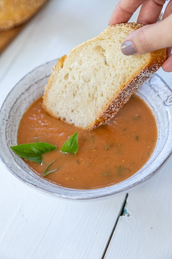 A bowl of tomato soup with a hand dipping a slice of bread in it.