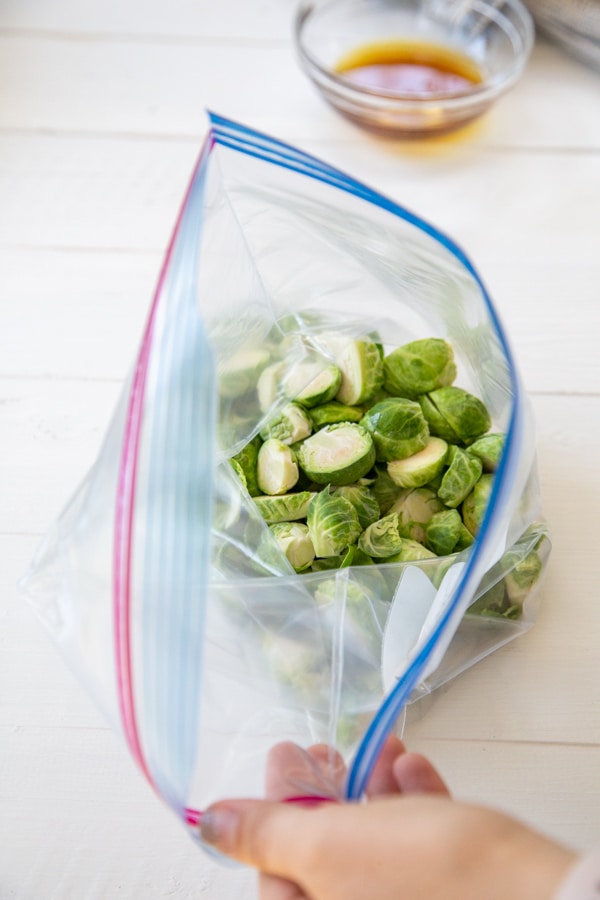 A hand holding a plastic bag filled with Brussels sprouts and a dish of maple syrup next to the bag.