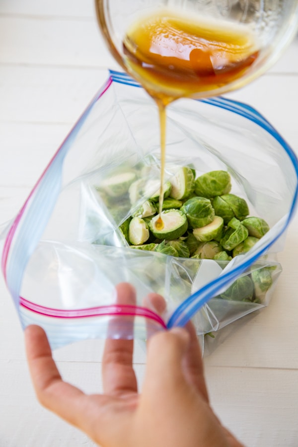 A hand holding a plastic bag with Brussels sprouts pouring maple syrup into the bag.