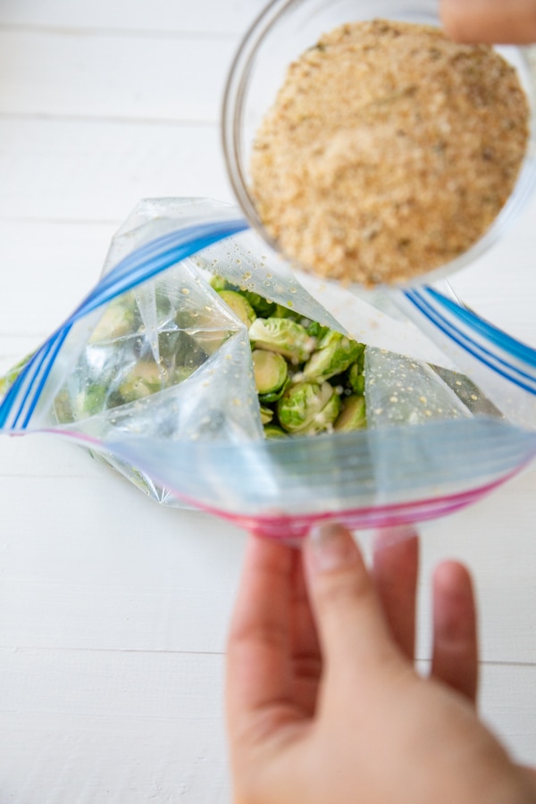 A hand pouring bread crumbs into a bag of Brussels sprouts.