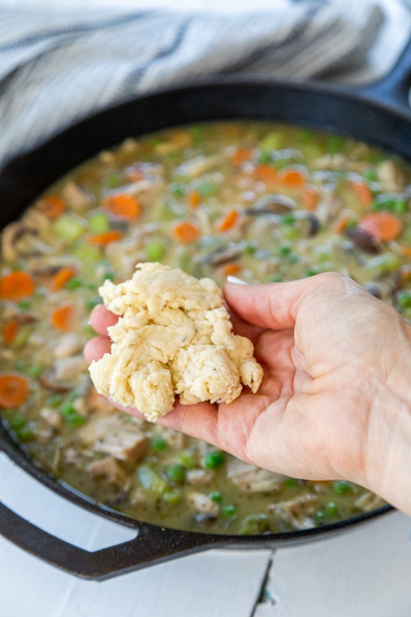 A hand holding a drop biscuit over chicken and vegetable stew in an iron skillet.