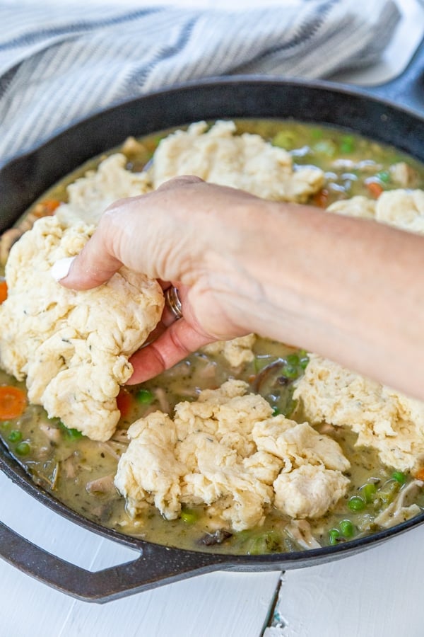 A hand placing drop biscuits over chicken and vegetable stew in an iron skillet.