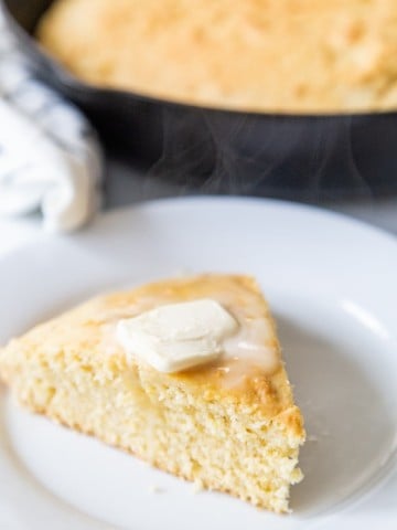 A slice of cornbread with butter on top on a white plate and a skillet of the cornbread behind it.