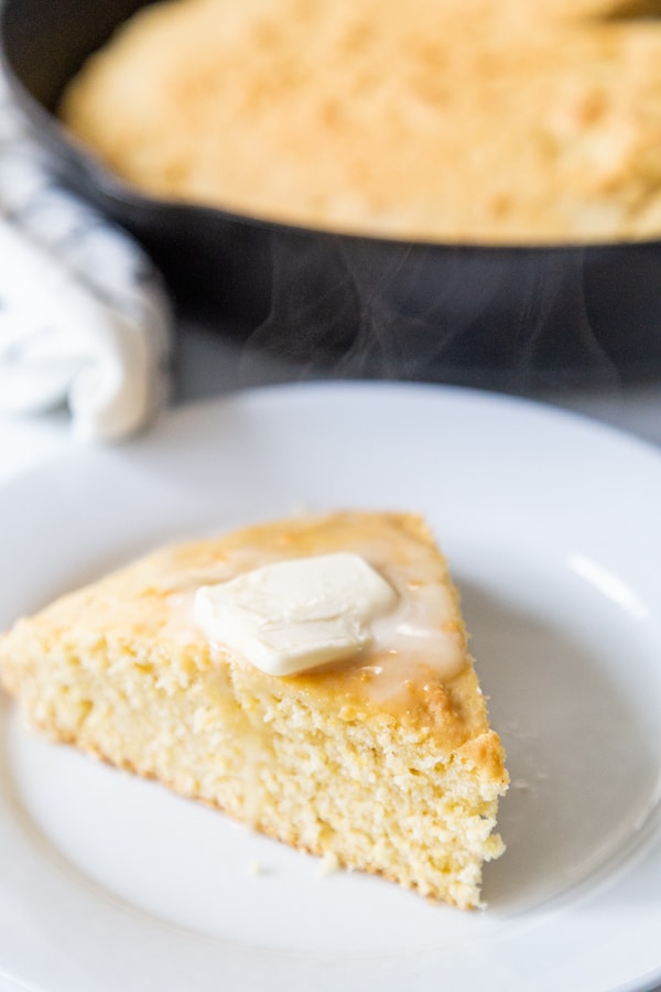 A slice of cornbread with butter on top on a white plate and a skillet of the cornbread behind it. 