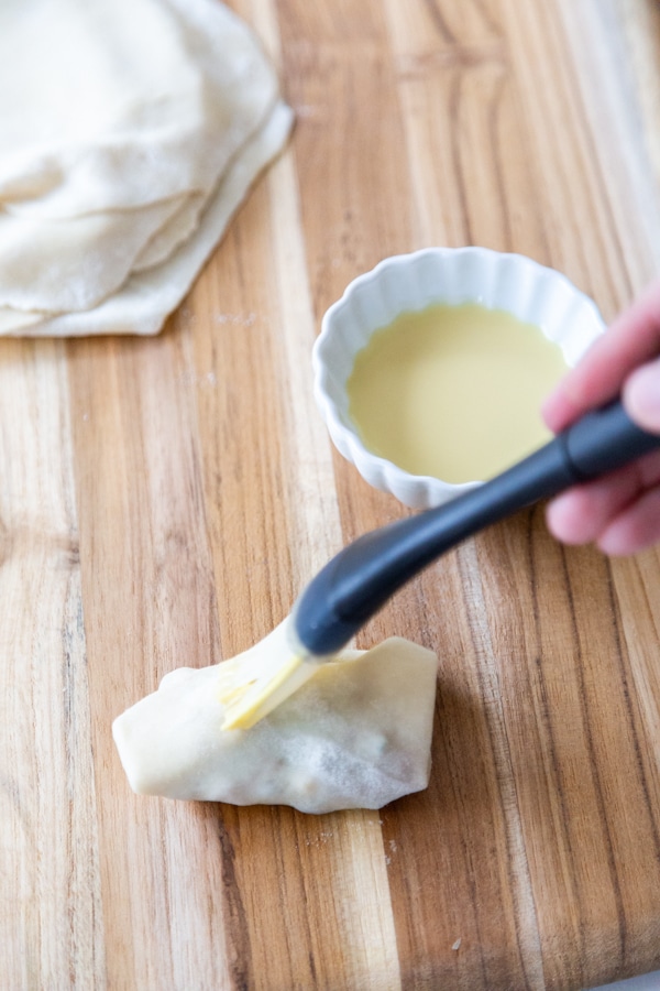 A hand brushing melted butter on an uncooked egg roll.