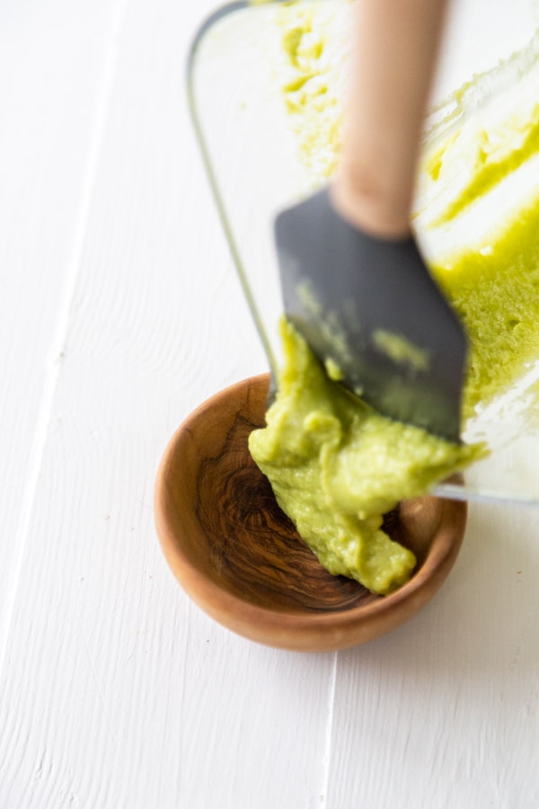 A hand scraping avocado dipping sauce out of a blender pitcher into a wood bowl. 