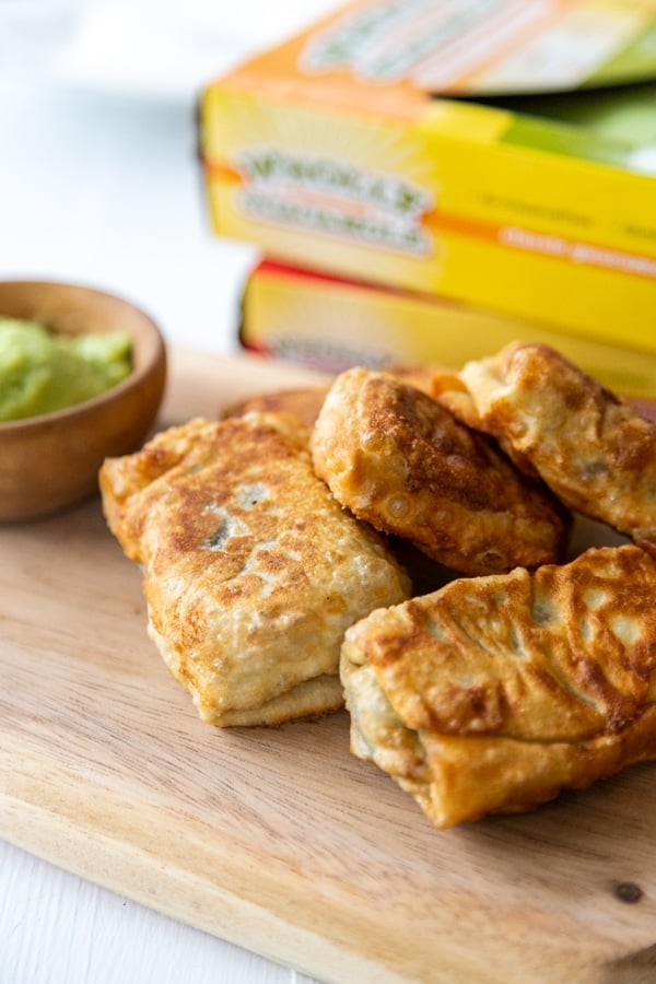 A stack of egg rolls on a wood board with a bowl of green dipping sauce and 2 boxes of guacamole in the background. 