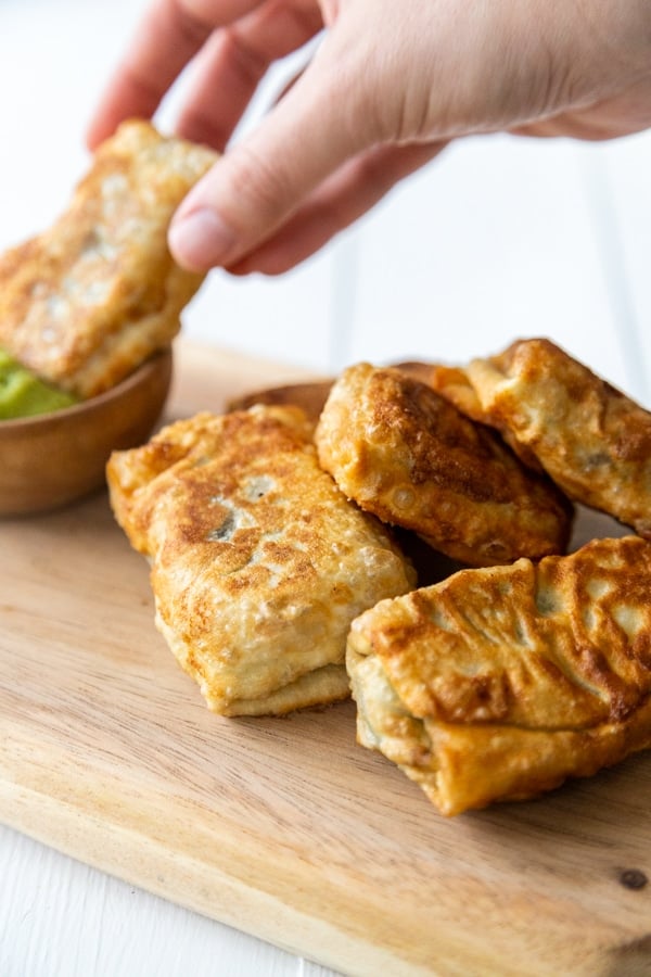 A hand dipping an egg roll into dipping sauce and a pile of egg rolls on a wood board. 