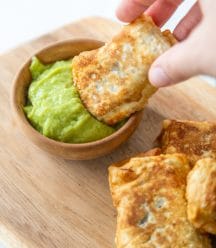 A hand dipping an egg roll into a wood bowl of green dipping sauce.