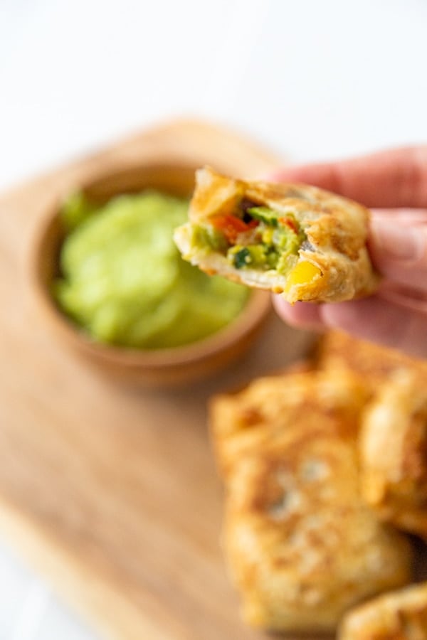 A hand holding an egg roll with a bite taken out of it over a wood board with more rolls and green sauce. 