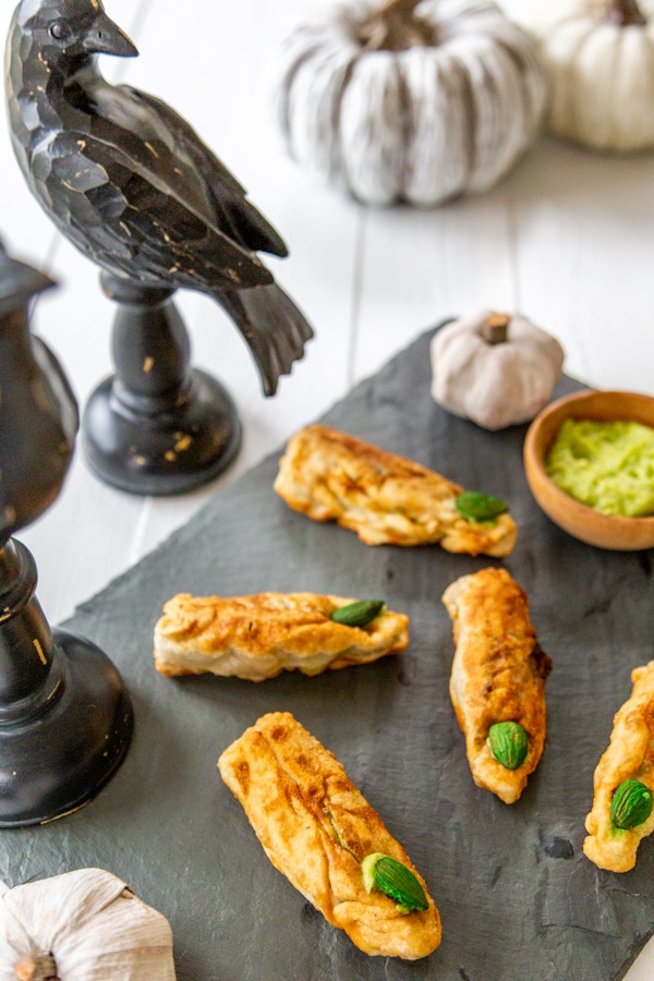 Finger shaped egg rolls on a slate board with wood crows and pumpkins next to it.