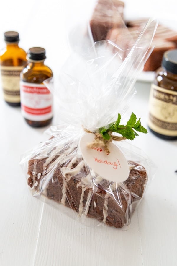 A mini chocolate cake loaf wrapped in cellophane and a ribbon with a sprig of mint, and Nielsen-Massey Vanillas extract bottles next to it. 