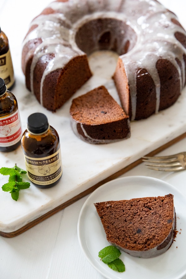 A chocolate bundt cake with icing drizzled over the top and a slice cut out of it, and brown bottles of extract next to it. 