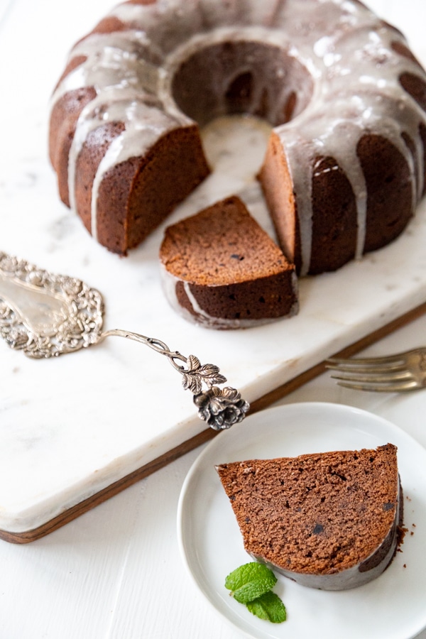A chocolate bundt cake with vanilla glaze drizzled down the top and a few slices cut out with a silver cake knife. 