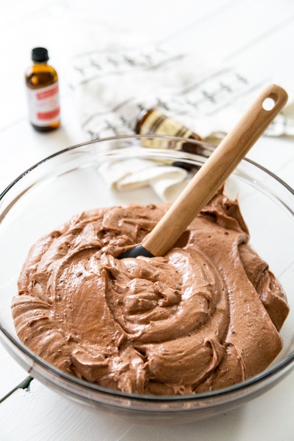 A clear glass mixing bowl with chocolate cake batter and a wooden spatula with bottles of Nielsen-Massey Vanillas extracts.