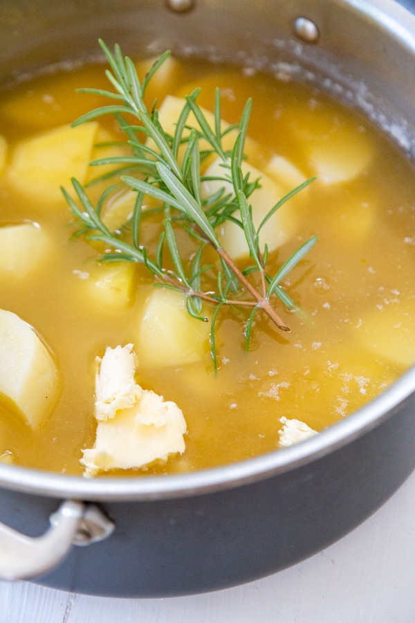 A pot of potatoes in broth with a clump of butter and rosemary sprigs.