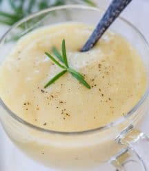A clear glass mug with potato soup and black pepper and a sprig of rosemary, and a spoon in the mug.