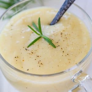 A clear glass mug with potato soup and black pepper and a sprig of rosemary, and a spoon in the mug.