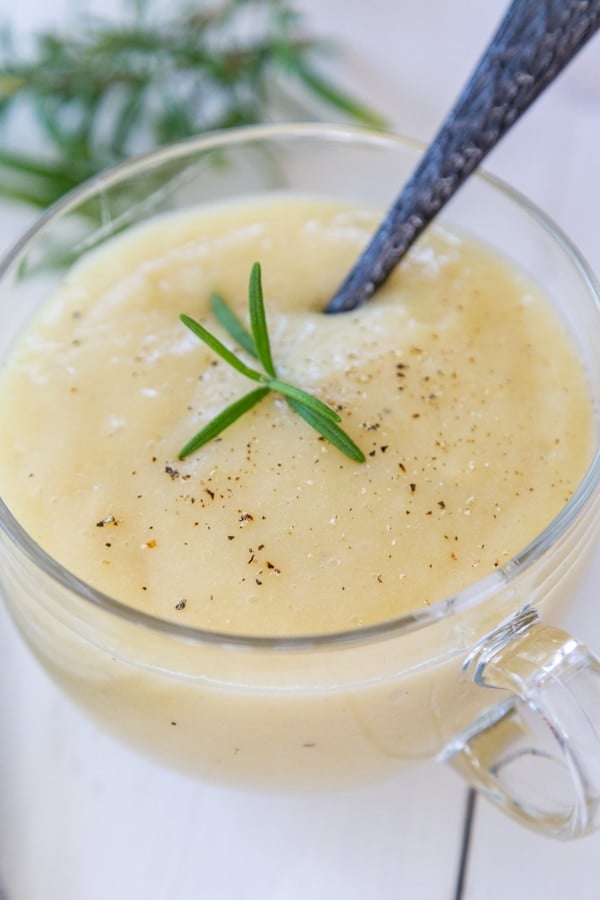 A clear glass mug with potato soup and black pepper and a sprig of rosemary, and a spoon in the mug.