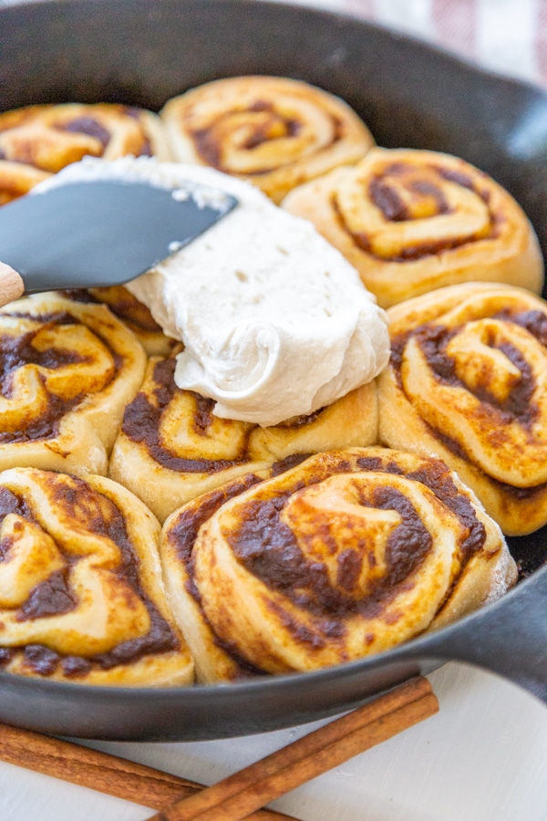 Cinnamon rolls with a spatula spreading cream cheese frosting over them. 