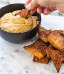 A hand dipping a pie crust chip into a black bowl of pumpkin dip.