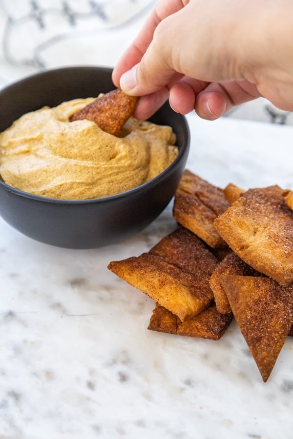 A hand dipping a pie crust chip into a black bowl of pumpkin dip.