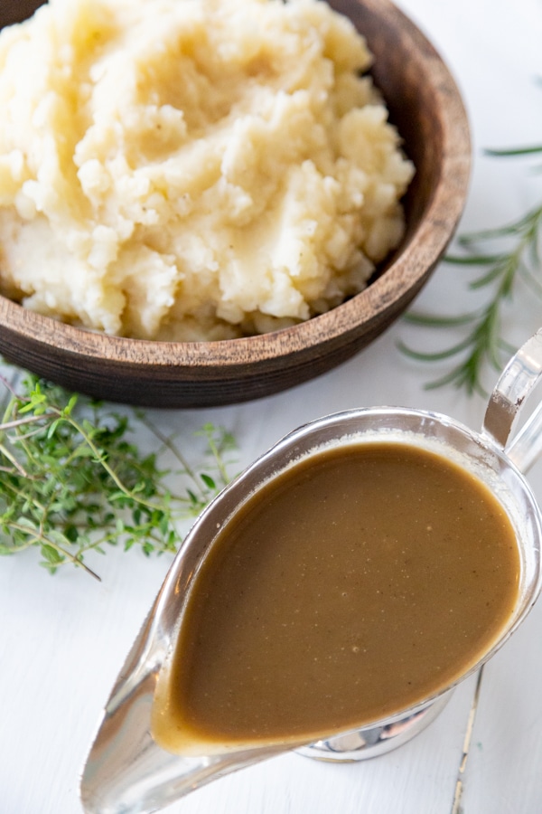 A wooden bowl with mashed potatoes and a silver gravy boat filled with brown gravy and fresh herbs on a white surface.