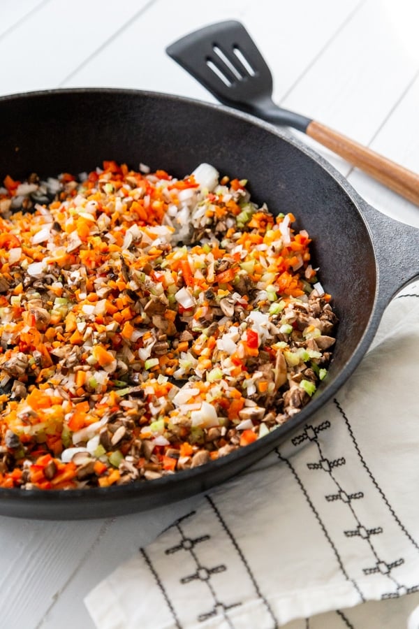 An iron skillet with seared veggies and a black and white towel and spatula next to the pan.