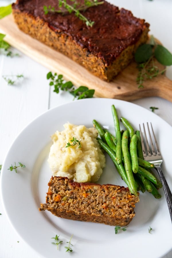 A white plate with a slice of lentil loaf, green beans, and mashed potatoes with the loaf in the background.