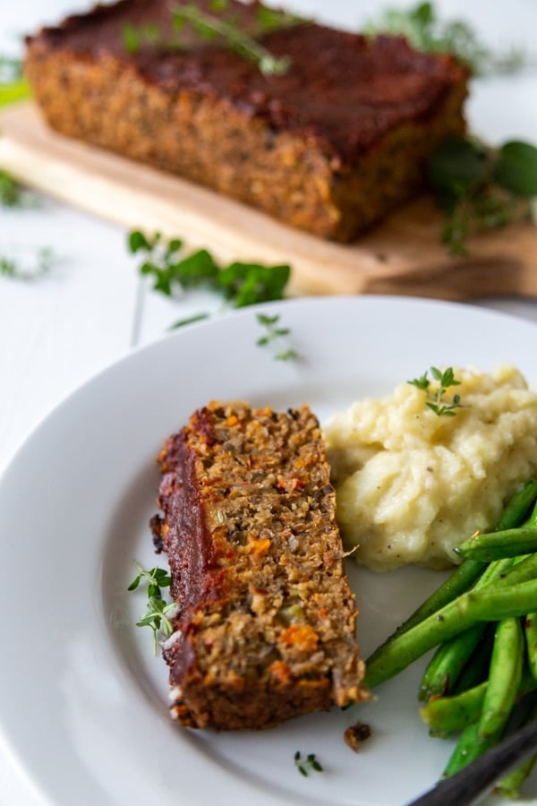 A slice of lentil loaf with green beans and mashed potatoes on a white plate with the whole loaf in the background.