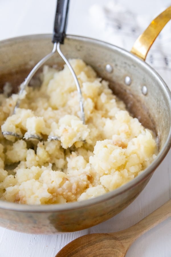 A potato masher mashing potatoes in a copper pan.