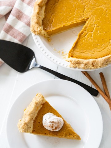 A slice of pumpkin pie with whipped topping on a white plate and the whole pie next to it.