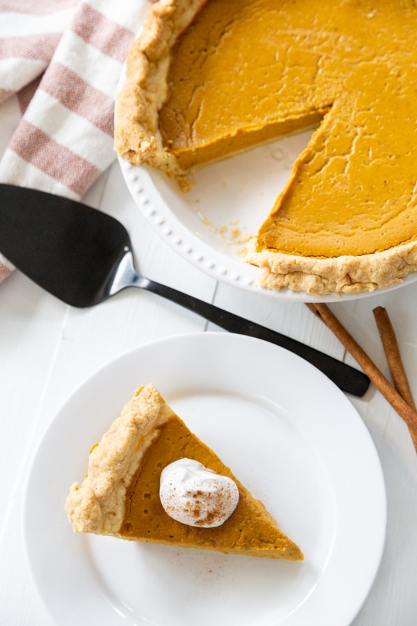A slice of pumpkin pie with whipped topping on a white plate and the whole pie next to it. 