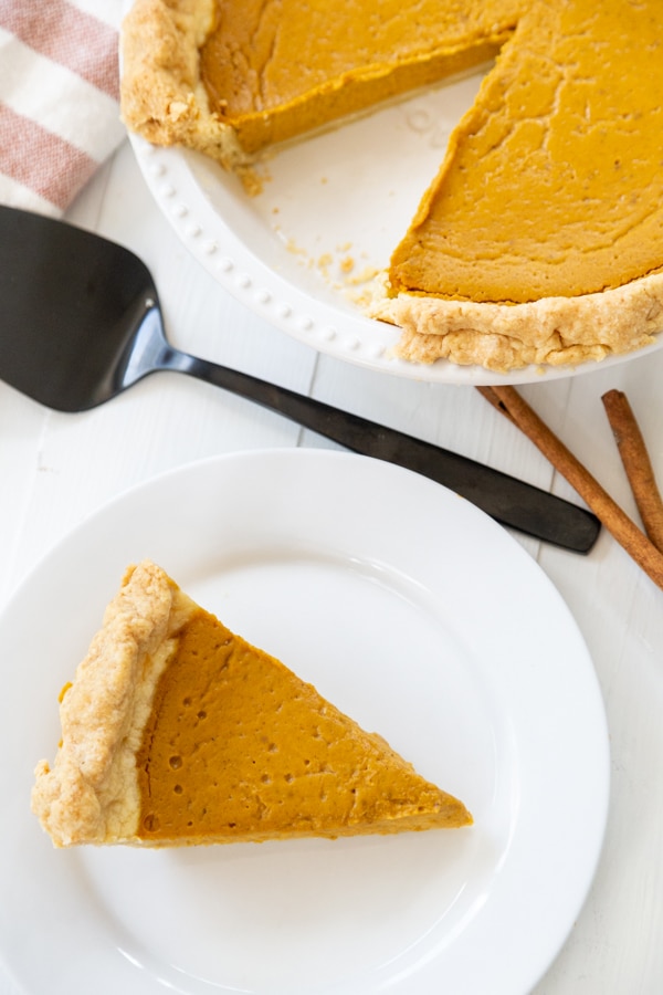 A slice of pumpkin pie on a white plate with the whole pie behind it. 