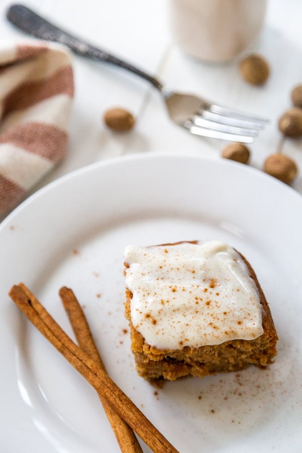 A slice of cake with cream cheese frosting on a white plate with 2 cinnamon sticks.