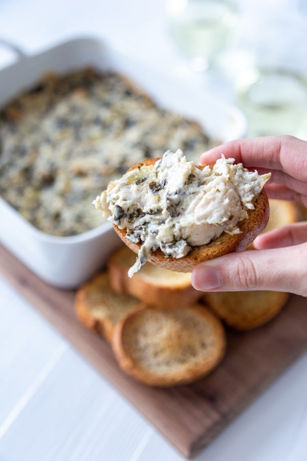 A hand holding toast with spinach artichoke dip spread on top, and a board with a pan of the dip and toast.