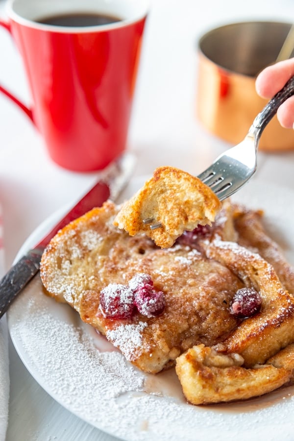 A fork full of French toast over a plate of French toast and a red mug of coffee. 
