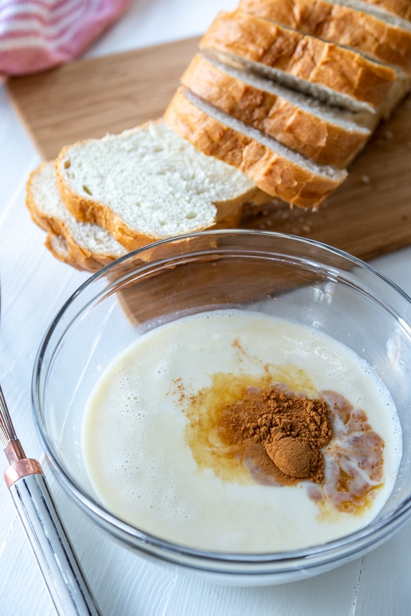 A glass bowl with milk and vanilla next to a board of sliced white bread.