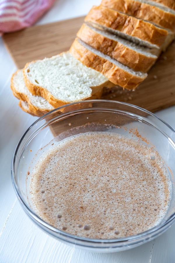 A glass bowl with milk and spiced next to a board of sliced white bread.