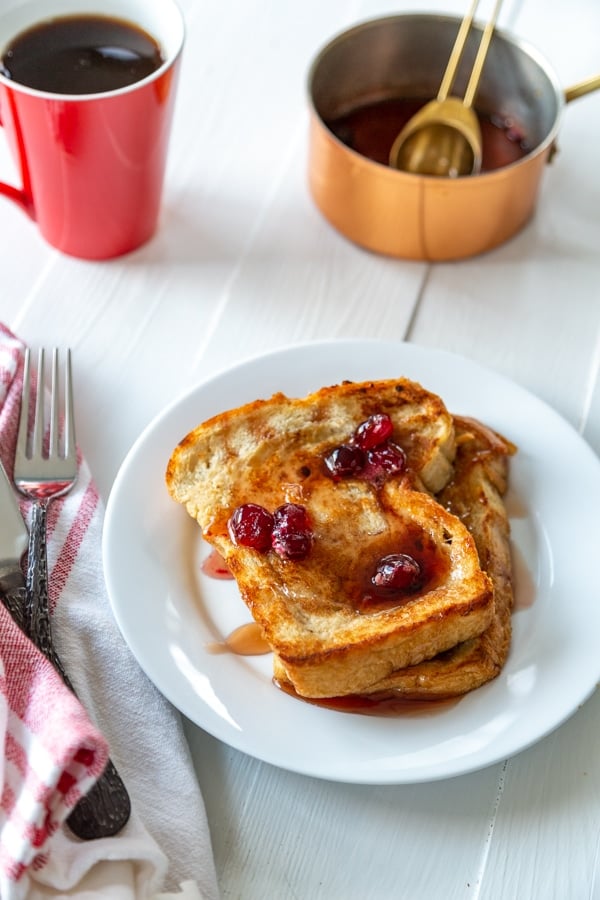 A plate of French toast with maple syrup and cranberries and a red cup of coffee and a copper pot of syrup. 