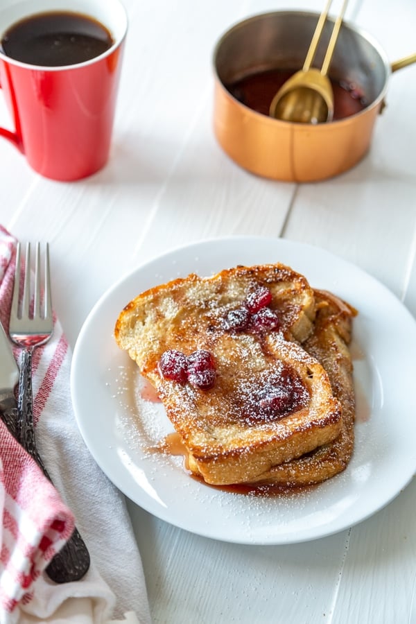 A white plate with 2 pieces of French toast with cranberry maple syrup and a copper pot with syrup and a red mug of coffee.