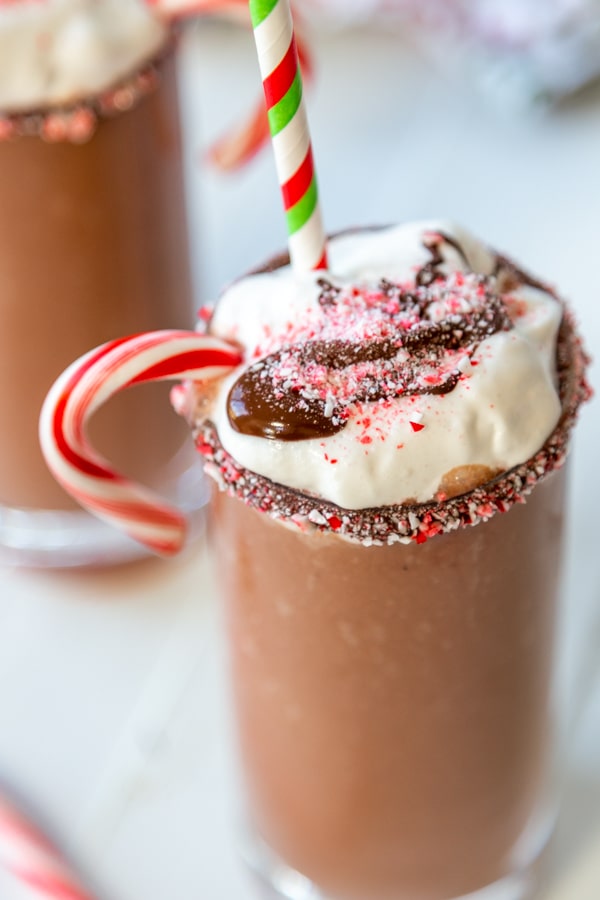 A frozen hot chocolate in a glass with a candy cane and whipped cream and melted chocolate. 