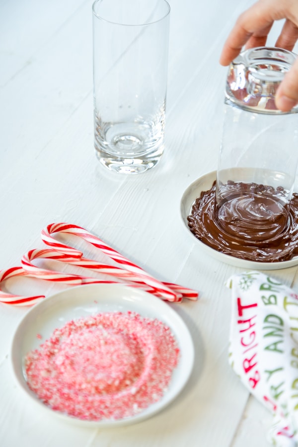 A hand dipping the rim of a glass in melted chocolate and a plate of ground candy cane. 