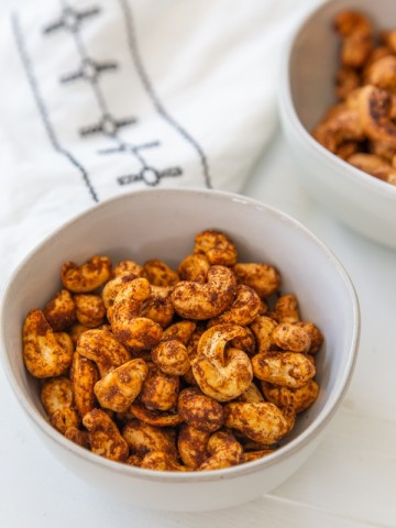 2 bowls of roasted spicy nuts and a white and black towel on a white table.