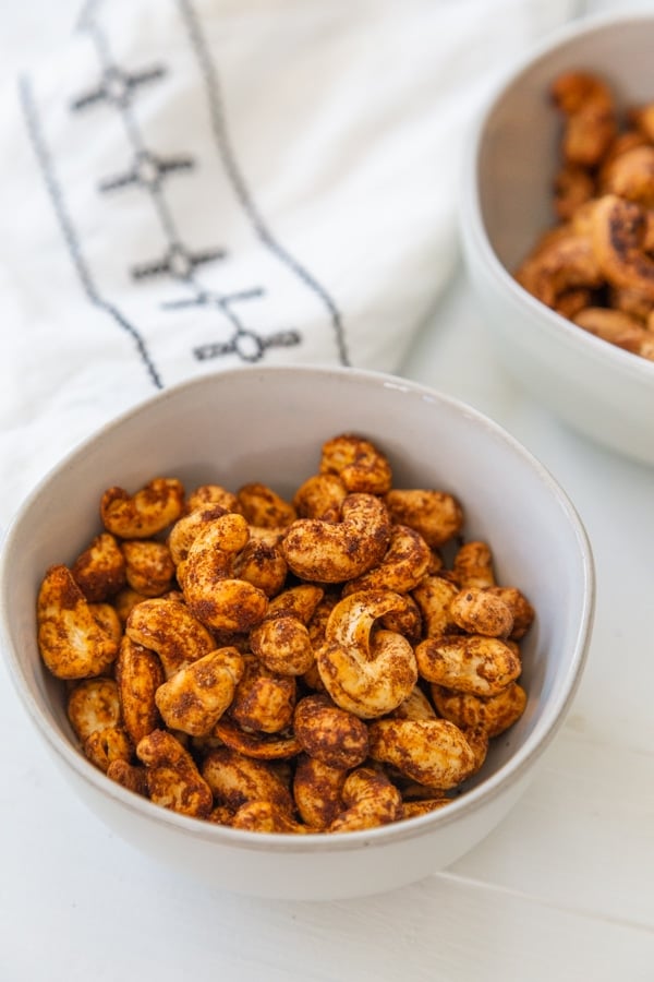 2 bowls of roasted spicy nuts and a white and black towel on a white table. 