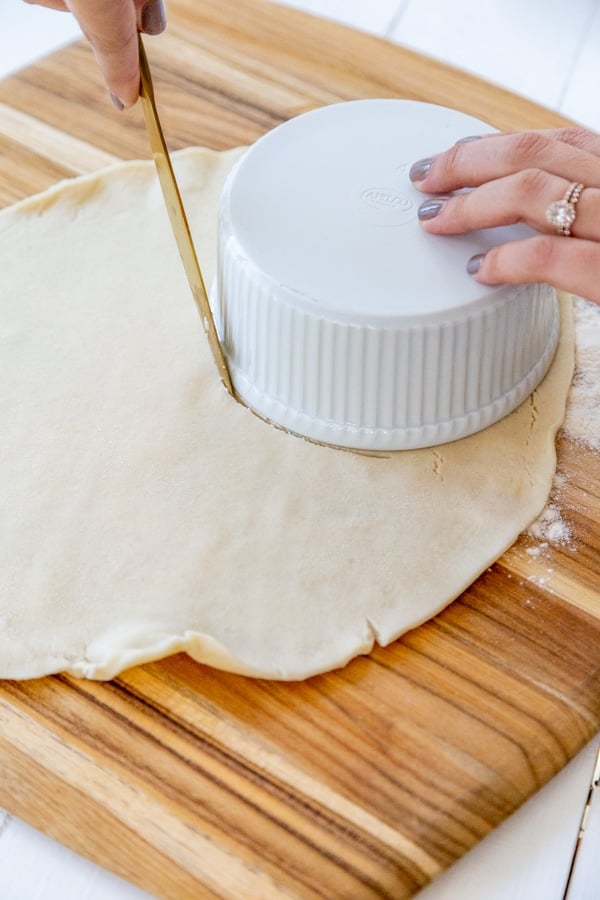 Pie dough rolled out on a wood board with a hand holding a bowl on the dough and cutting around it to make a circle.
