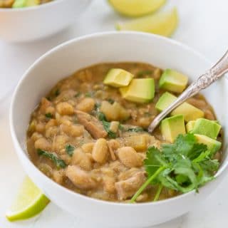 A bowl of white bean chili with avocado and cilantro on top and a spoon in the bowl.