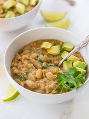 A bowl of white bean chili with avocado and cilantro on top and a spoon in the bowl.