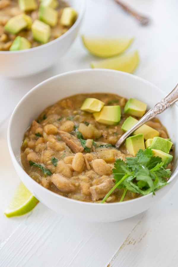 A bowl of white bean chili with avocado and cilantro on top and a spoon in the bowl.